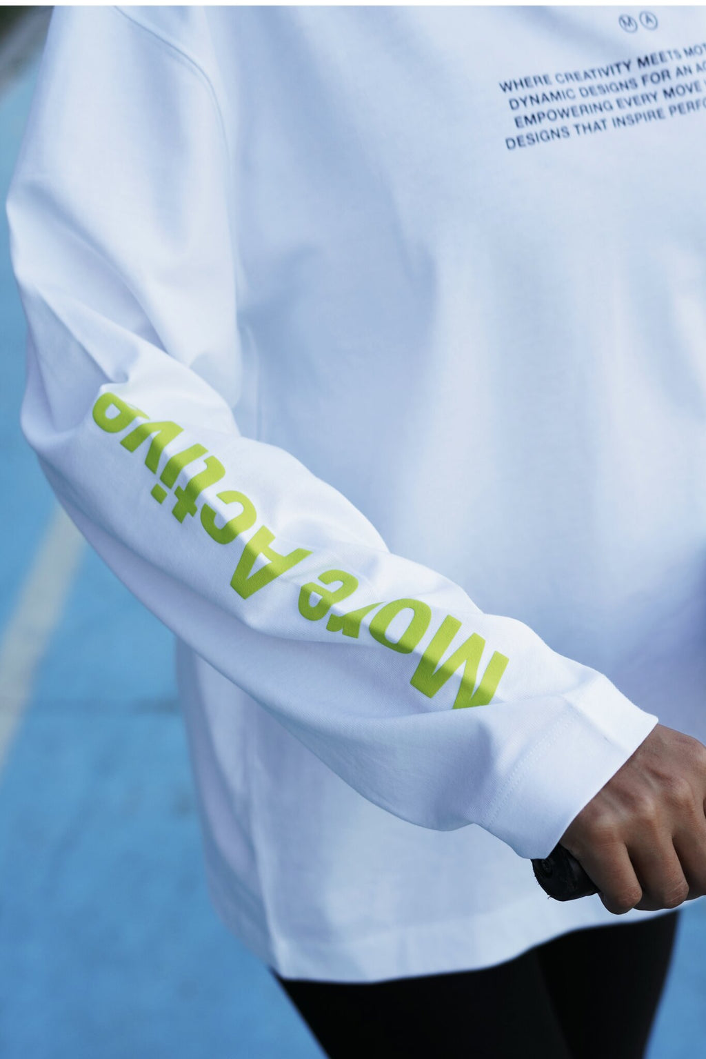A close-up of a white long-sleeve athletic top worn by a person on a blue court. The right sleeve has large lime green text reading 'More Active', and the chest has small dark text starting with 'WHERE CREATIVITY MEETS.