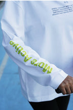A close-up of a white long-sleeve athletic top worn by a person on a blue court. The right sleeve has large lime green text reading 'More Active', and the chest has small dark text starting with 'WHERE CREATIVITY MEETS.
