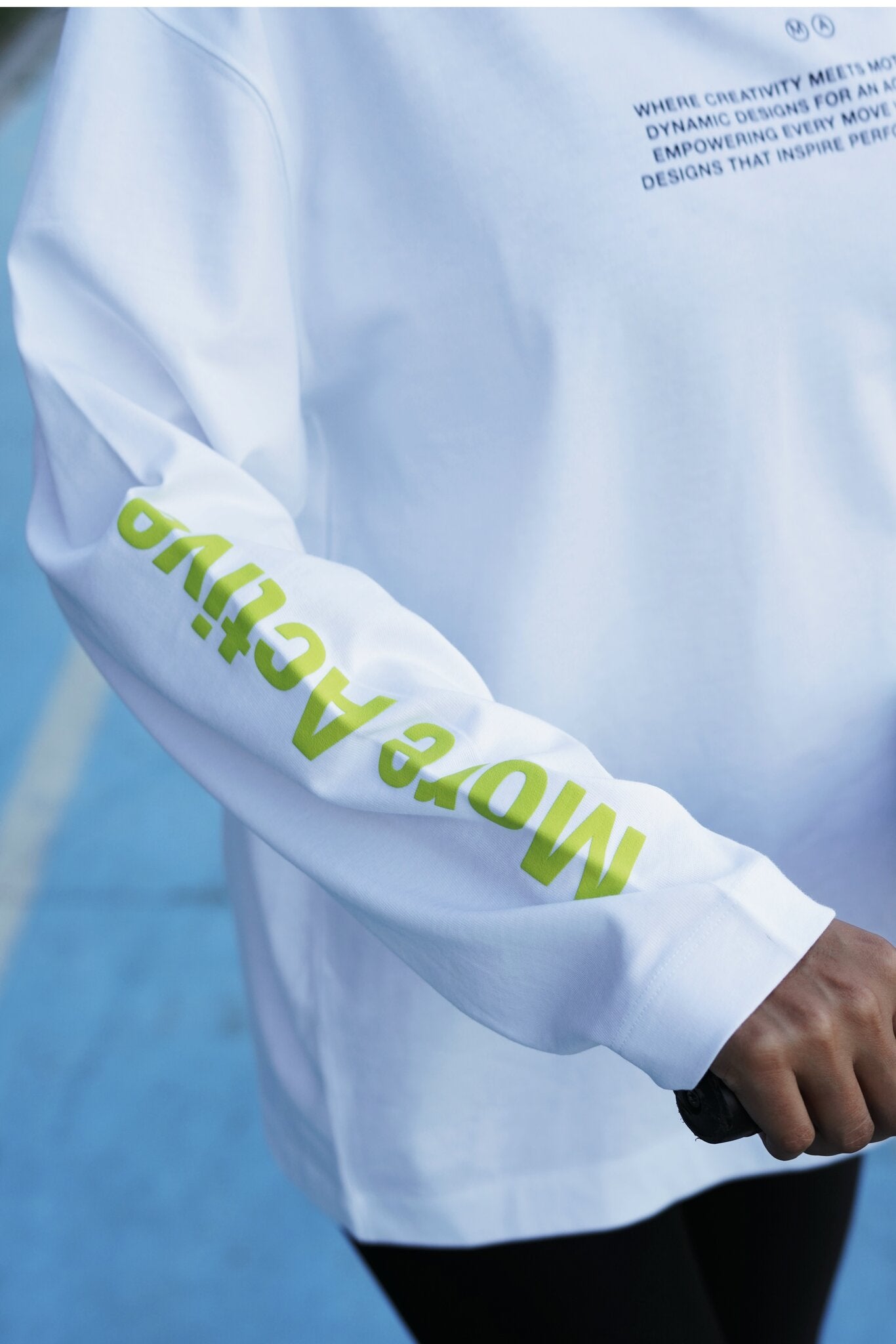 A close-up of a white long-sleeve athletic top worn by a person on a blue court. The right sleeve has large lime green text reading 'More Active', and the chest has small dark text starting with 'WHERE CREATIVITY MEETS.