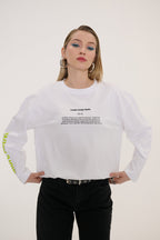 A woman modeling a white long-sleeve t-shirt with black text graphics on the chest and sleeve, paired with black jeans, against a plain off-white background.