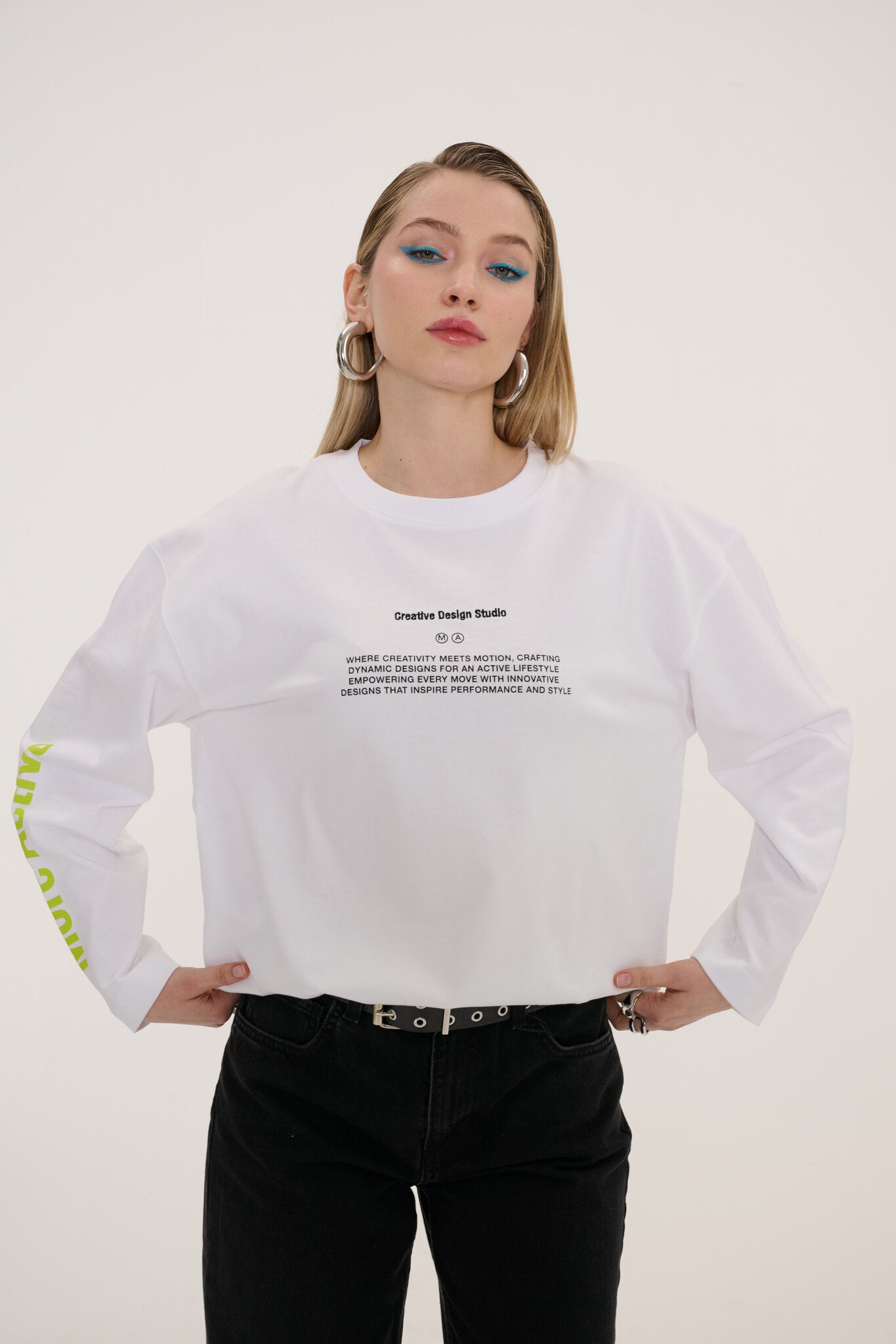 A woman modeling a white long-sleeve t-shirt with black text graphics on the chest and sleeve, paired with black jeans, against a plain off-white background.