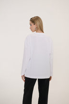 A woman shown from the back wearing a white long-sleeve top and black pants, standing against a plain off-white studio background.